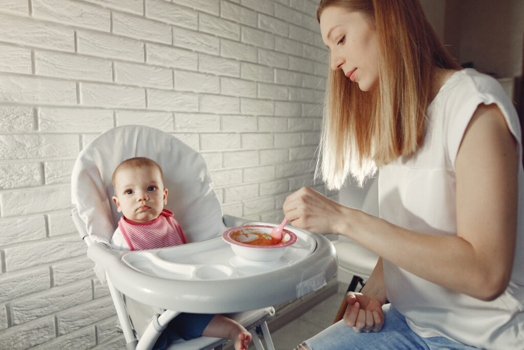 Mother introducing solids to her baby in a high chair as part of a baby-fed and spoon-fed approach.
