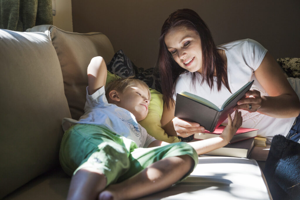 A smiling mother leans over her young son on a sunlit couch, holding an open book as he lies back on a pillow and follows along with the story.