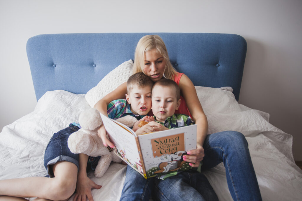 A mother in a pink top and jeans sits against a blue headboard on a white bedspread, holding an open picture book as her two young sons snuggle close—one clutching a teddy bear—while they all look at the illustrations together.