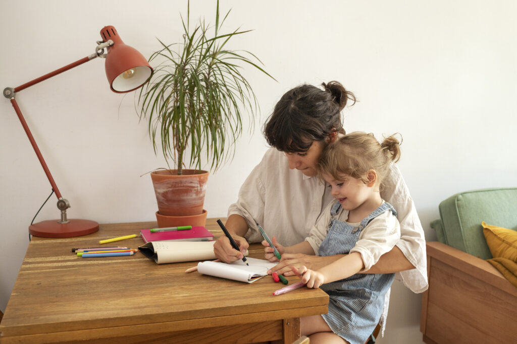 A mother and her little daughter sit at a wooden desk covered in markers and a sketchpad; the mother guides her daughter’s hand as they draw together under a red desk lamp beside a potted plan